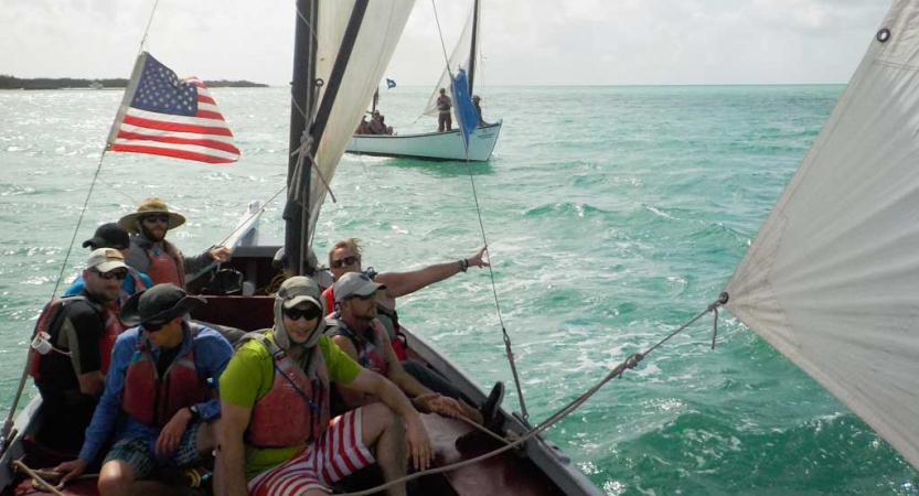 A group of people wearing life jackets sit in a sailboat flying an American flag. 